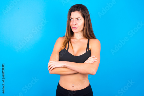 Displeased Young beautiful sportswoman doing sport wearing sportswear over blue background with bad attitude, arms crossed looking sideways. Negative human emotion facial expression feelings.