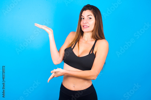 Young beautiful sportswoman doing sport wearing sportswear over blue background pointing aside with both hands showing something strange and saying: I don't know what is this. Advertisement concept.