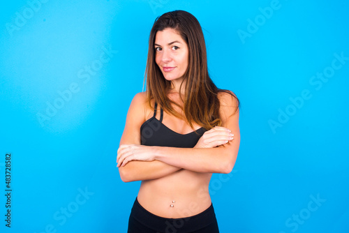 Waist up shot of self confident Young beautiful sportswoman doing sport wearing sportswear over blue background has broad smile, crosses arms, happy to meet with colleagues.