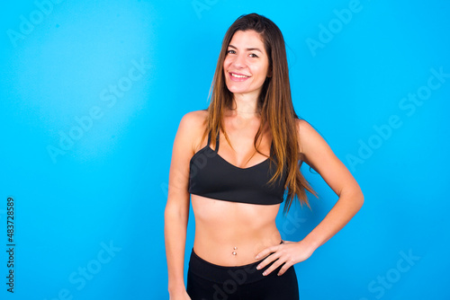 Studio shot of cheerful Young beautiful sportswoman doing sport wearing sportswear over blue background keeps hand on hip, smiles broadly.