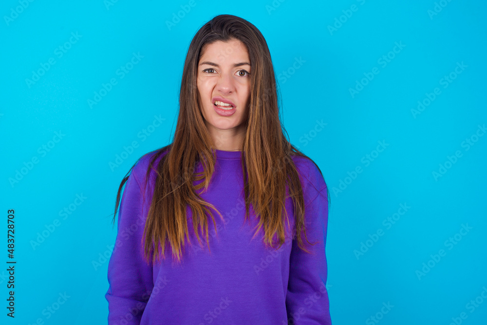 Fototapeta premium Portrait of dissatisfied young beautiful caucasian woman wearing purple sweater against blue smirks face, purses lips and looks with annoyance at camera, discontent hearing something unpleasant