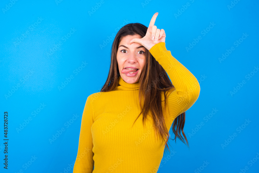 young beautiful caucasian woman wearing yellow turtleneck sweater against blue background gestures with finger on forehead makes loser gesture makes fun of people shows tongue young beautiful caucasian woman wearing yellow turtleneck sweater against blue background gestures with finger on forehead makes loser gesture makes fun of people shows tongue