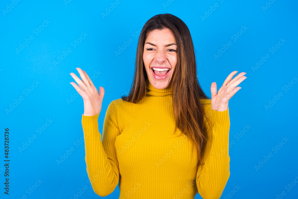 Fototapeta premium Crazy outraged young beautiful caucasian woman wearing yellow turtleneck sweater against blue background screams loudly and gestures angrily yells furiously. Negative human emotions feelings concept