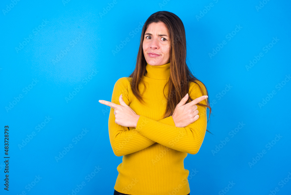 Serious young woman wearing yellow turtleneck sweater against blue background crosses hands and points at different sides hesitates between two items. Hard decision concept Serious young woman wearing yellow turtleneck sweater against blue background crosses hands and points at different sides hesitates between two items. Hard decision concept