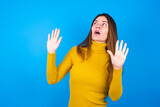 young beautiful caucasian woman wearing yellow turtleneck sweater against blue keeps palms forward and looks with fright above on ceiling tries to defense herself from invisible danger opens mouth.