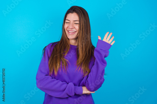 Overjoyed successful oung beautiful caucasian woman wearing purple sweater against blue background raises palm and closes eyes in joy being entertained by friends