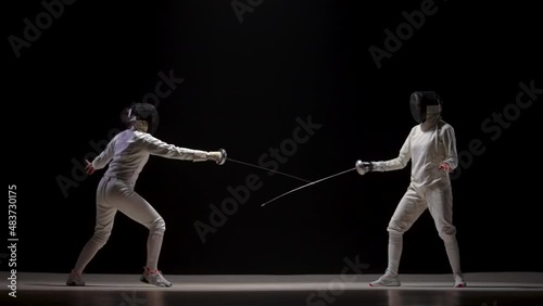 Two female athletes train on foil at fencing tournament. Young female swordsmen in white uniforms and fencing masks fight duel against black background. Fencing sports school. Slow motion. photo