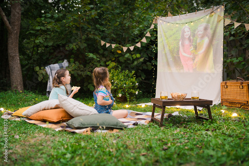 Family summer outdoor movie night. Girls sitting on blanket and pillows, eating homemade popcorn and watching film on DIY screen with from projector. Summer outdoor weekend activities with kids.