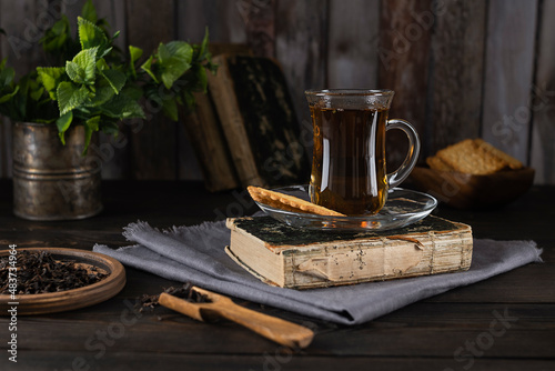 Tea in a glass cup on an old book on a light gray napkin. Mint leaves in a tin mug and cookies in a wooden bowl in the background. Wooden background
