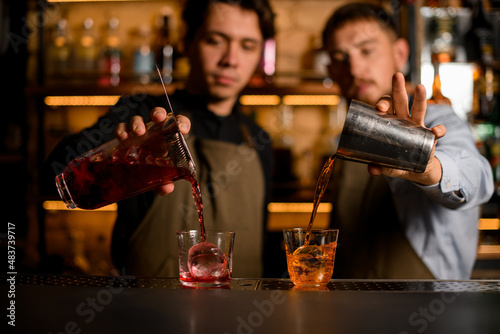 Two glasses stands on bar and two bartenders fills them with alcohol drinks
