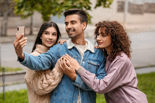 Man with two beautiful women taking selfie outdoors. Polyamory concept