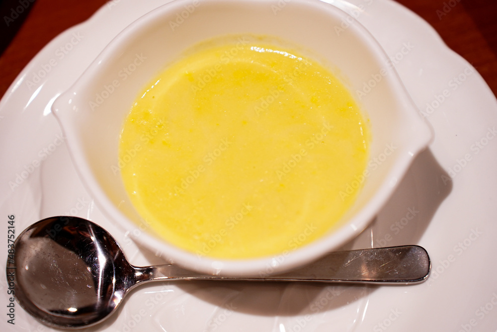 A stylish small white bowl of sweet corn soup made of brand corn from Hokkaido Japan with a round metal spoon and a white plate underneath A stylish small white bowl of sweet corn soup made of brand corn from Hokkaido Japan with a round metal spoon and a white plate underneath