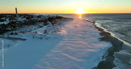 Snow on beach. Waves crash on shore at sunrise. Lighthouse beacon. Descending aerial of snow-covered dunes.