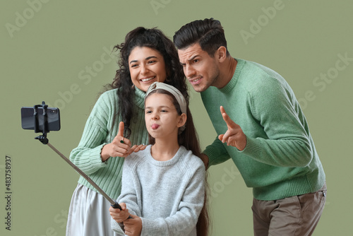 Happy parents with little daughter in warm sweaters taking selfie on green background