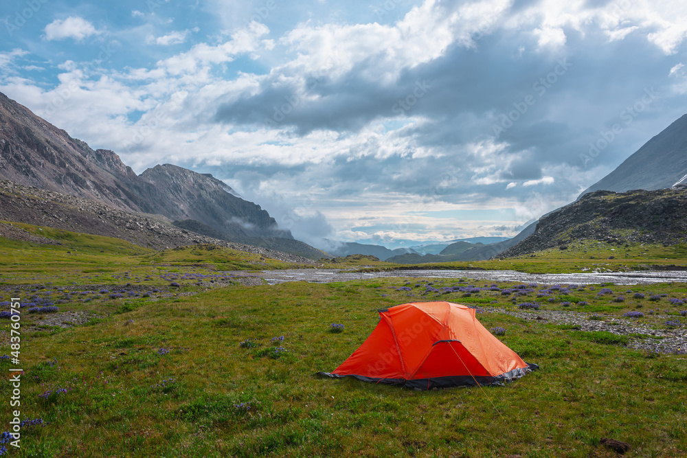 Scenic mountain landscape with tent in rain drops in sunlight under cloudy sky. Dramatic alpine scenery with vivid orange tent on grass among purple flowers in green mountain valley in rainy weather.