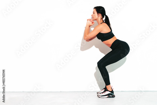 Portrait of fitness confident woman in black sports clothing. Sexy young beautiful model with perfect body. Female isolated on white wall in studio. Stretching out before training. Making squats