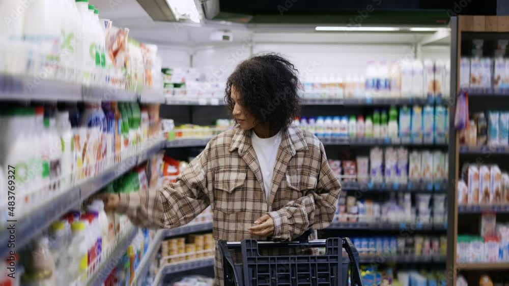 Positive black woman with mobile device and trolley purchasing products at mall