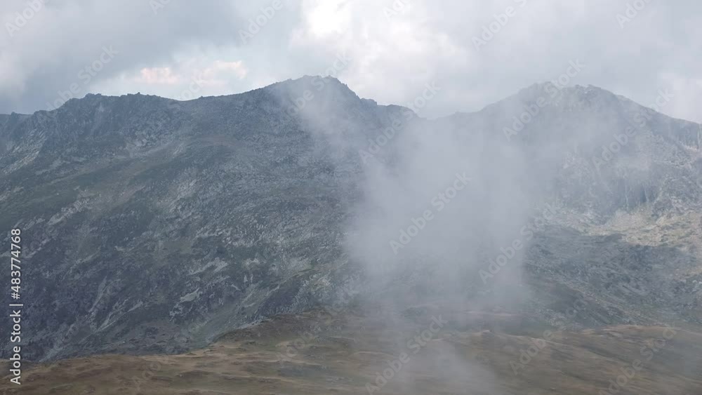 View from Rila mountain peak on impressive steep rocky cliffs under a moody sky and mist View from Rila mountain peak on impressive steep rocky cliffs under a moody sky and mist
