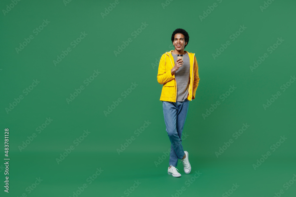 Full size young black curly man 20s years old wears yellow waterproof raincoat outerwear hold takeaway delivery craft paper brown cup coffee to go isolated on plain green background studio portrait. Full size young black curly man 20s years old wears yellow waterproof raincoat outerwear hold takeaway delivery craft paper brown cup coffee to go isolated on plain green background studio portrait.