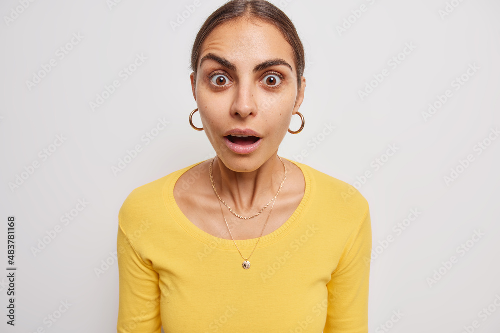 Amazed beautiful young woman with dark combed hair keeps mouth opened says wow and stares impressed at camera poses against white background drops jaw from big news. Human reactions concept. Amazed beautiful young woman with dark combed hair keeps mouth opened says wow and stares impressed at camera poses against white background drops jaw from big news. Human reactions concept.