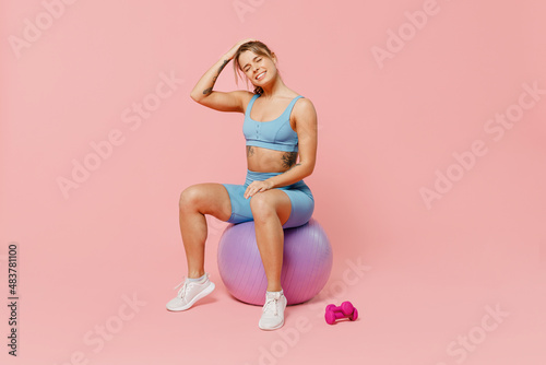 Full body young sporty fitness trainer instructor woman wear blue tracksuit spend time in home gym sit on fitball hold neck suffer from pain isolated on plain pink background. Workout sport concept.