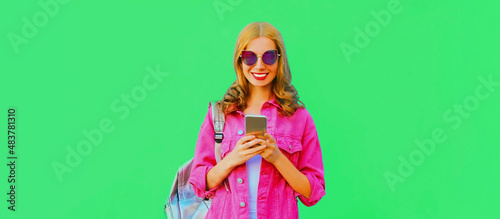 Portrait of stylish happy smiling young woman with smartphone wearing pink jacket, sunglasses on green background