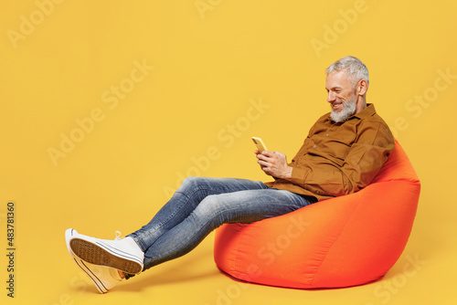Full size body length happy fun elderly gray-haired bearded man 40s years old wears brown shirt sit in bag chair hold in hand use mobile cell phone isolated on plain yellow background studio portrait. photo