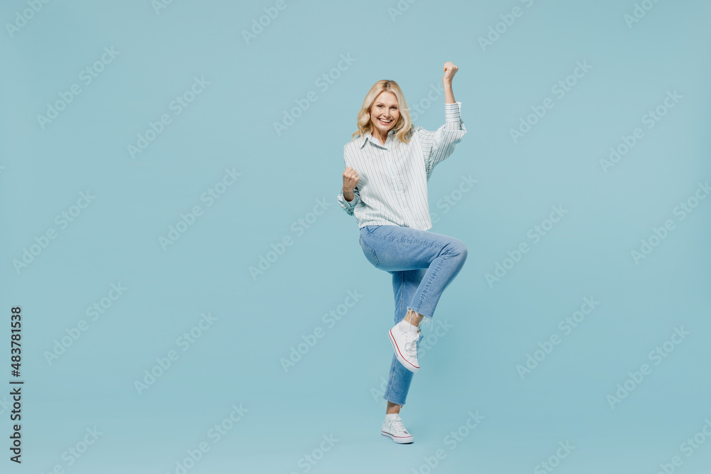 Full body overjoyed elderly woman 50s in casual striped shirt do winner gesture clench fist raise up leg isolated on plain pastel light blue color background studio portrait. People lifestyle concept. Full body overjoyed elderly woman 50s in casual striped shirt do winner gesture clench fist raise up leg isolated on plain pastel light blue color background studio portrait. People lifestyle concept.