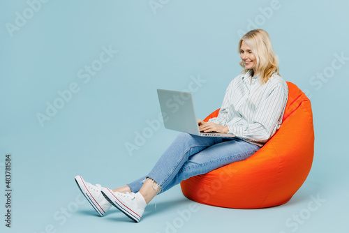 Full body elderly woman 50s in casual striped shirt sit in bag chair hold use work on laptop pc computer isolated on plain pastel light blue color background studio portrait. People lifestyle concept.