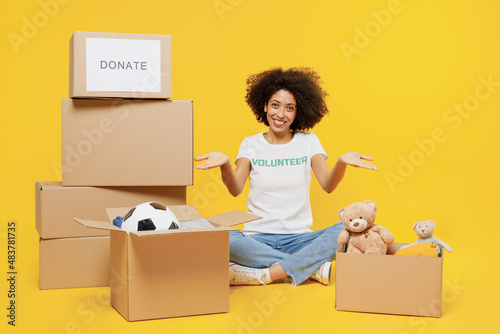 Full body young woman of African American ethnicity wears white volunteer t-shirt sit near boxes show presents isolated on plain yellow background. Voluntary free work assistance help grace concept. photo