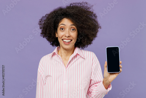 Young smiling happy woman of African American ethnicity 20s in pink striped shirt hold in hand use mobile cell phone with blank screen workspace area isolated on plain pastel light purple background