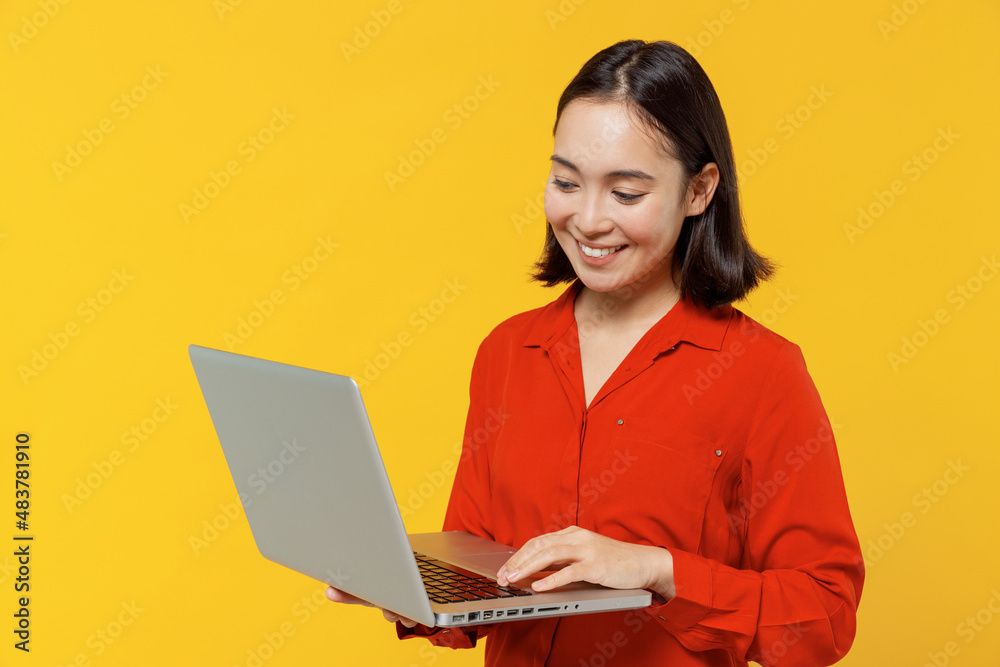 Businesslike smiling young woman of Asian ethnicity 20s years old wears orange shirt hold use work on laptop pc computer typing searching chatting isolated on plain yellow background studio portrait. Businesslike smiling young woman of Asian ethnicity 20s years old wears orange shirt hold use work on laptop pc computer typing searching chatting isolated on plain yellow background studio portrait.