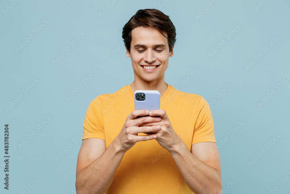 Young smiling man 20s wear yellow t-shirt hold in hand use mobile cell phone chatting browsing read message isolated on plain pastel light blue background studio portrait. People lifestyle concept Young smiling man 20s wear yellow t-shirt hold in hand use mobile cell phone chatting browsing read message isolated on plain pastel light blue background studio portrait. People lifestyle concept