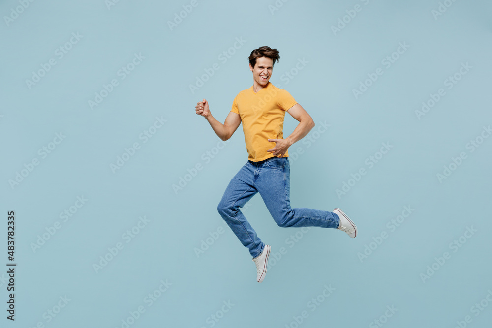 Full body musician happy caucasian brunette young man 20s wearing yellow t-shirt jump high play guitar isolated on plain pastel light blue color background studio portrait. People lifestyle concept.