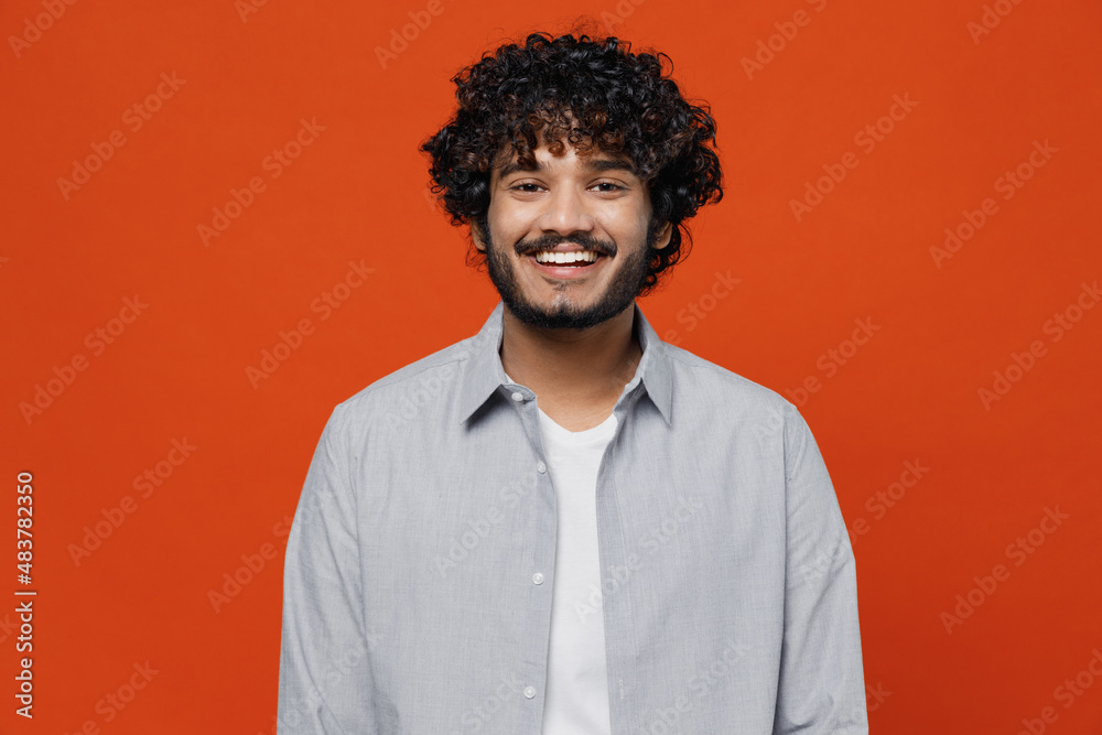 Smiling fascinating cheerful blithesome young bearded Indian man 20s years old wears blue shirt looking camera isolated on plain orange background studio portrait. People emotions lifestyle concept. Smiling fascinating cheerful blithesome young bearded Indian man 20s years old wears blue shirt looking camera isolated on plain orange background studio portrait. People emotions lifestyle concept.