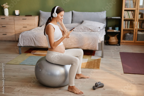 Young pregnant woman in headphones using mobile phone to listen to music while exercising on fitness ball in the bedroom photo