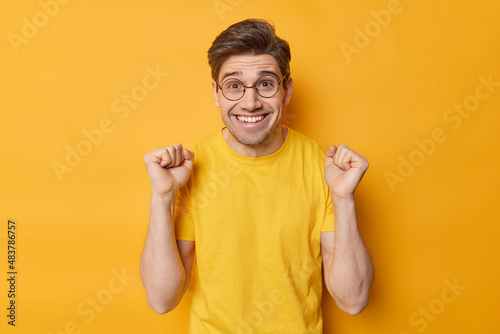 Happy positive man cheers over something clenches fists and rejoices victory smiles toothily feels very glad wears round spectacles and casual t shirt isolated over yellow background. Triumph and luck