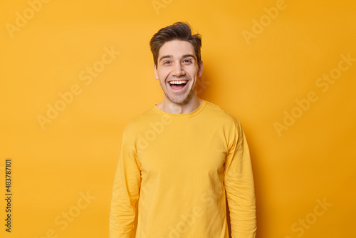 Horizontal shot of happy young European man laughs joyfully expresses positive emotions dressed in casual jumper isolated over vivid yellow background. Monochrome shot. Positive guy indoors.
