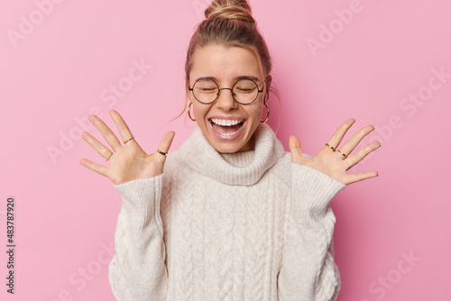 Fotobehang Horizontal shot of happy woman with combed hair raises palms wears round spectac