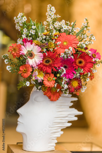 Colorful bouquet decorative gerber flowers in a white ceramic vase in the shape of a head, standing on the windowsill