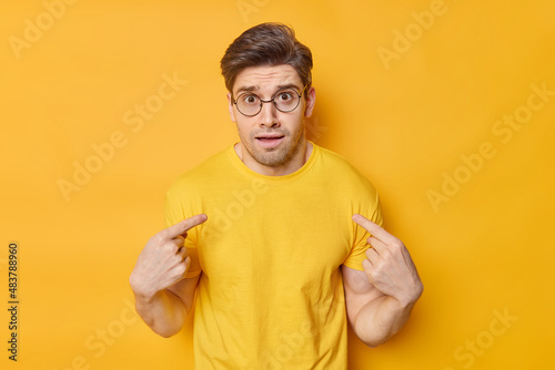Tableau sur toile Indignant young man looks puzzled points at himself feels embarrassed wears transparent round spectacles and casual t shirt asks who me isolated over vivid yellow background