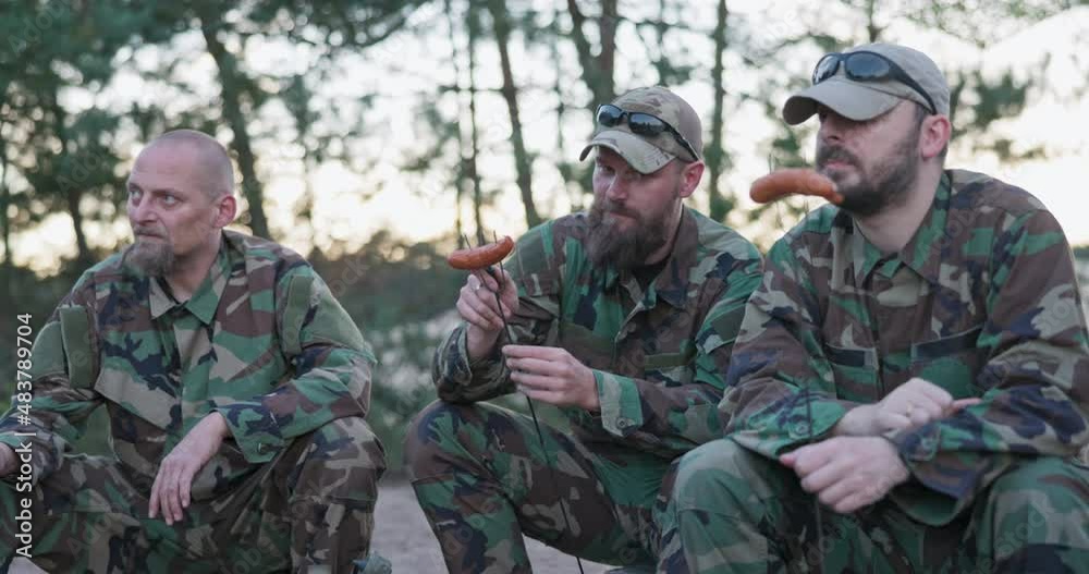 Military men in moro uniforms resting after an afternoon shift at base in field sitting on crates outside preparing campfire scooping sausage onto sticks guy with a beard cuts meat with a penknife