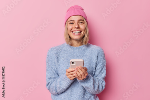 Happy fair haired female technology user holds smartphone glad to chat online with boyfriend wears hat and jumper poses against pink studio background. Cellular technology and networking concept © wayhome.studio 