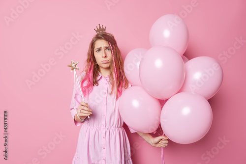 Unhappy frustrated woman comes on party poses with inflated balloons and magic wand wears small princess crown and dress poses against pink background. People holidays and celebration concept