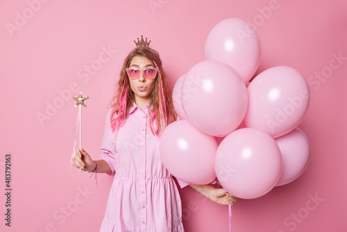 Impressed astonished fashionable woman wears trendy sunglasses dress and crown on head has image of princess holds bunch of inflated helium balloons magic wand keeps lips rounded poses indoor