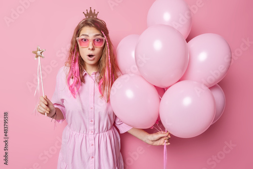 Impressed stunned European woman wears trendy sunglasses and dress reacts on shocking newsholds bunch of inflated balloons and magic wand isolated over pink background. Holiday event concept