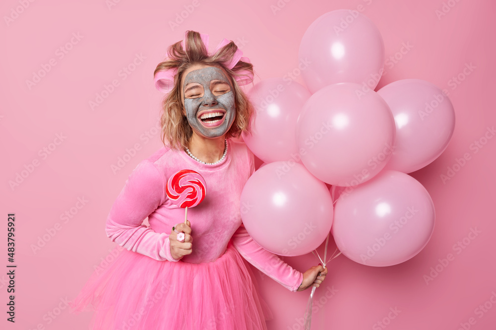 Fototapeta premium Glad happy woman applies beauty clay mask on face giggles positively holds sweet candy on stick and bunch of helium balloons wears festive dress makes hairstyle with hair rollers enjoys celebration