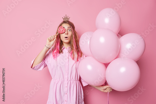 Impressed astonished long haired woman covers eyes with heart shaped candy on stick wears dress and crown holds bunch of inflated balloons comes on birthday party isolated over pink background