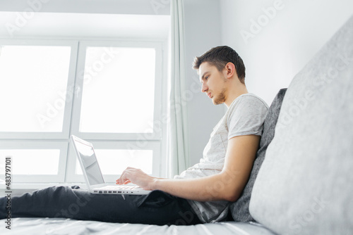 Young attractive smiling guy is browsing, working online on his laptop while sitting at home on a cozy sofa at home