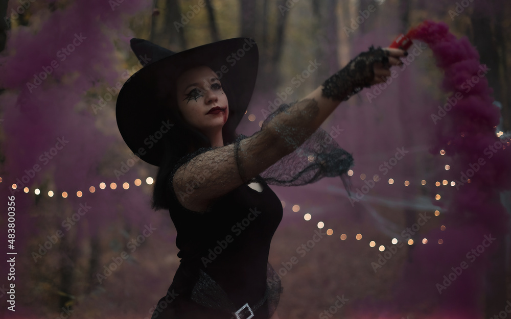 Portrait in the woods with a sexy woman with a hat on her head and a black dress, made up with fake blood. She holds a colorful fumigant in her hand.
